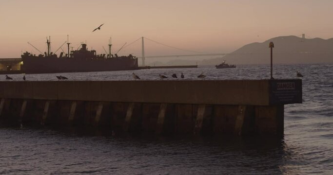 City port view of a water jetty in foreground, a big ship and bridge in background, at sunset
