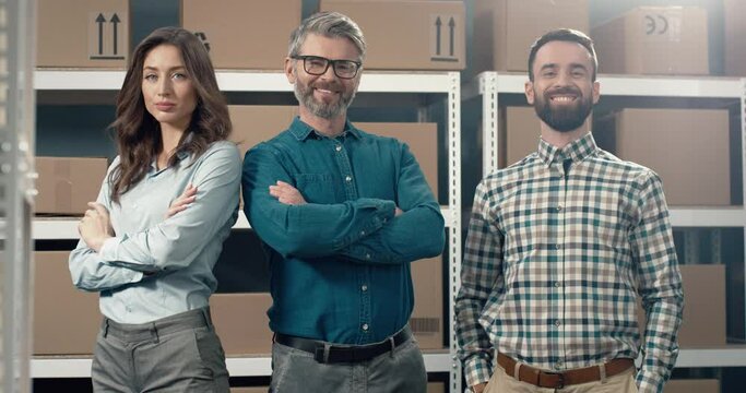 Portrait of three successful colleagues logistics warehouse managers standing on cardboard boxes background with arms crossed looking at camera smiling.