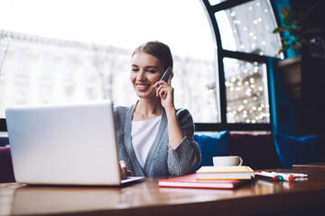 Positive female entrepreneur talking on smartphone while browsing laptop