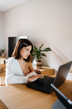 Young mom freelancer breastfeeding baby and working on laptop remotely