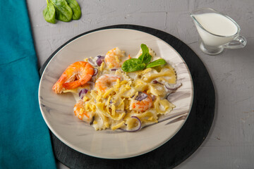 Farfalle pasta with shrimps in a creamy sauce on a gray plate on a concrete background next to a blue napkin and a jug of cream.