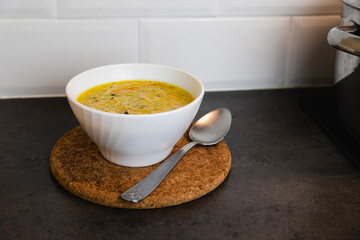 Small glass bowl full of chicken soup with spoon on the side and wooden table protector on the kitchen counter.