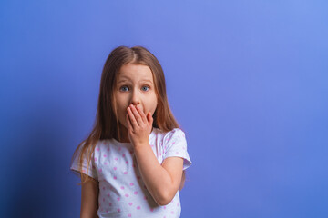 a portrait of a surprised, shocked little girl, covering her mouth with her hand, stands on a purple background. the child doesn't want to talk. Keeps a secret, Mystery learned