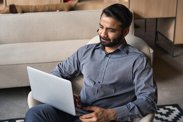 Serious bearded indian business man working on laptop from home office sitting in chair. Male...