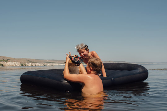 Two boys swim on an inflatable mattress with Corgi dog in googles