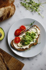avocado toast from whole grain bread with cream cheese tomatoes and microgreen. sandwich on a plate on a white marble surface. vegan breakfast