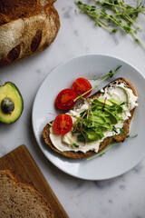avocado toast from whole grain bread with cream cheese tomatoes and microgreen. sandwich on a plate on a white marble surface. vegan breakfast