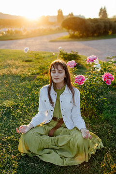 Young female sitting on lawn with blooming peony and doing yoga