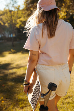 Curvy blonde girl on her back carrying her skateboard. She wears a pink t-shirt, white shorts and a cap.