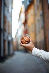 a man's hand in a white sweater holds a cinnamon bun on a blurred background of the old city....