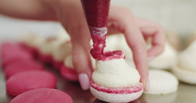 Beautiful pink macaroons fill with pink jam on a pink background. Nature pastry macaroons. Shot of hands of female pastry chef holding white macaron with ganache and squeezing red fruit jam from bag