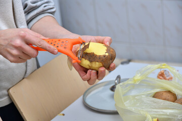 Hands of a young woman peeling potatoes with kitchen utensil on a wooden board
