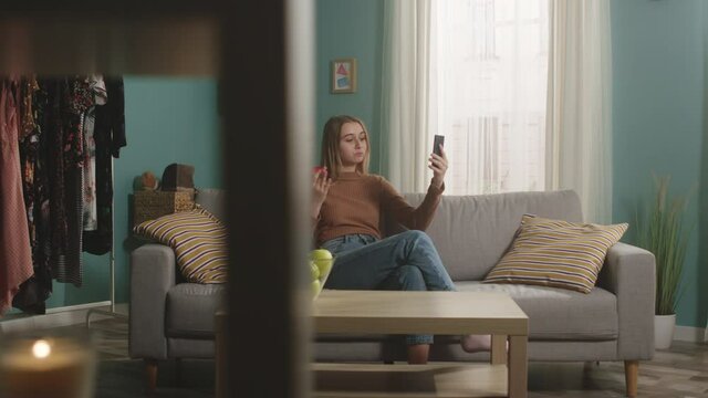Young girl in blue jeans and brown sweater sits on beige sofa on window background, bites red apple and talks on video call. On coffee table is glass jar with green apples. Camera dollies horizontally