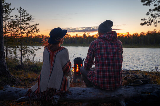 A girl and a guy are sitting near a fire and drinking tea on a background of a pine forest