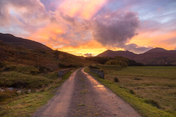 Evening glow sunset in the Scottish highland with rural scene of a single track gravel road, a stone brigde leading into the woods and mountains