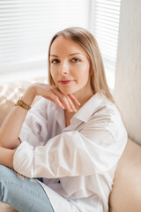 Naklejka premium A beautiful blonde girl in a white oversized shirt and blue jeans sits in a bright sunny room on a beige sofa
