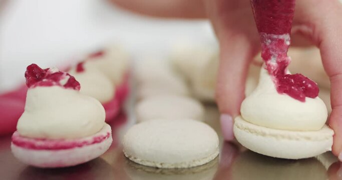Making Caramel Macarons. Close up cropped shot of hands of female confectioner, squeezing red fruit jam into the ganache cream on macarons shells. Filling macarons with ganache cream and fruit jam