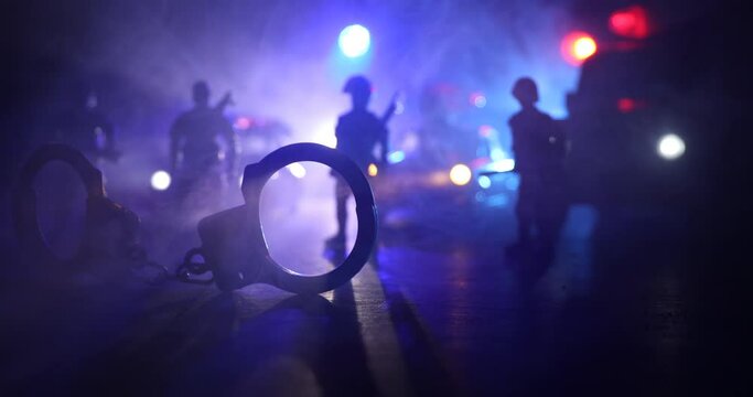 Police raid at night and you are under arrest concept. Silhouette of handcuffs with police car on backside. Image with the flashing red and blue police lights at foggy background. Selective focus