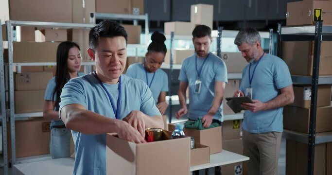 Multi-ethnic team of volunteers collaborating in workshop. Chinese male adult worker collecting food supply free delivery box smiling into camera.