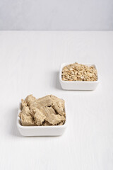 Halva, also halvah or halwa asian homemade sweet dessert served in square plate together with its main ingredient sunflower seeds on white wooden table. Vertical image. Cooking concept