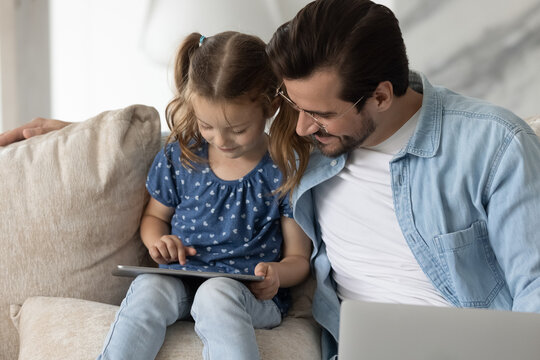 Loving young Caucasian father and little daughter relax on couch have fun using playing online on tablet. Happy caring dad and small girl child talk speak on webcam call on pad. Technology concept.