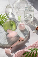 Obraz na płótnie Canvas Gua Sha massager and pink quarz face roller on marble stone background. Face care products, glass crystal ball and exotic palm leaves. Towel,moisturizing cream, skincare tonic, essential oil, serum.