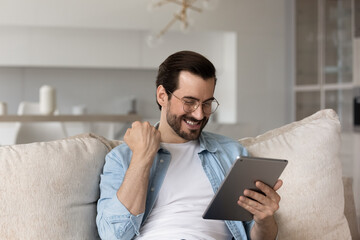 Overjoyed young Caucasian man look at tablet triumph get pleasant good email or message online....