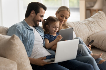Smiling young family with little 7s daughter sit on sofa at home use electronic gadgets together. Happy Caucasian parents relax with small girl child have fun with devices. Technology concept.