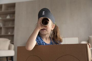 Close up of little Caucasian girl have fun play pirate game in living room on weekend. Small child in hat look in pipe spyglass engaged in funny playful activity at home. Entertainment concept.