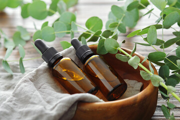 Two bottles of eucalyptus essential oil in wooden bowl and eucalyptus twigs on table. - obrazy, fototapety, plakaty