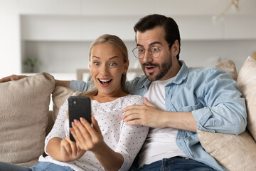 Stunned young man and woman user look at smartphone screen shocked by unexpected news online....