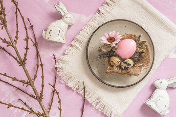 Handmade Easter breakfast decoration: jute fabric, pink Easter egg, natural quail eggs and feathers on a rustic plate, pinkish background, top view