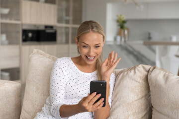 Happy millennial Caucasian woman sit on sofa at home look at cellphone wave talk speak on video call online. Smiling young female have digital webcam conference on smartphone. Virtual event concept.