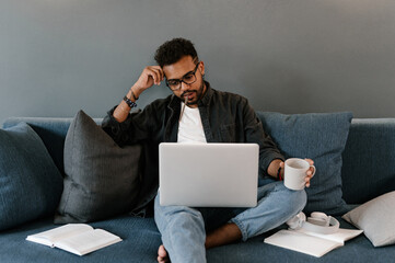 Thoughtful ethnic student doing homework on sofa