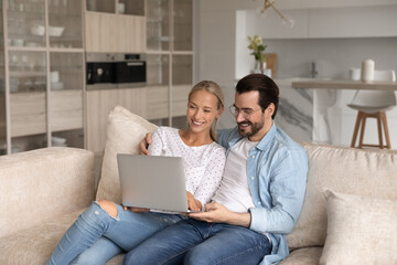 Overjoyed young Caucasian couple renters relax in modern renovated apartment using computer. Happy millennial man and woman rest on couch talk on video call on laptop at home. Rental concept.