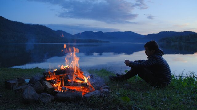 Teenage boy sitting on the lake with a smartphone, silhouette shooting at sunset.