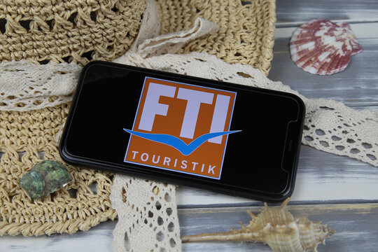 Viersen, Germany - March 1. 2021: Closeup of smartphone with logo lettering of fti touristik group travel agency with sun hat and shells on wood table