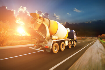 Concrete mixer truck on a countryside road in motion with trees against a sky with a sunset