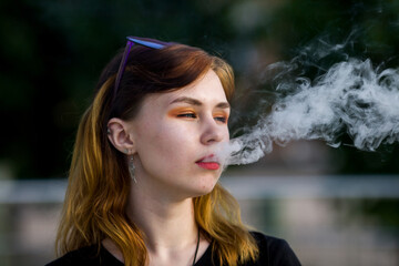 Naklejka premium Vape teenager. Young pretty white caucasian girl with glasses on her head an electronic cigarette on the street in the summer. Deadly bad habit.