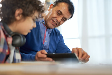 Happy latin middle aged father watching his son, school boy doing his homework during remote...