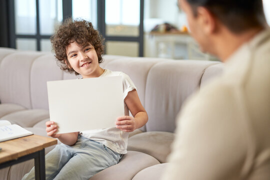 Happy latin boy showing the drawing to his dad while spending time with his father, playing in the living room at home