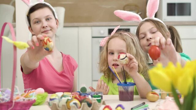 Three beautiful girls show drawings on Easter eggs in front of the camera. There is an Easter basket on the wooden table. Easter holiday. 4k video.