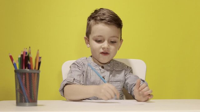 Smiling little boy at the table draw with crayons for mum on yellow background. Creativity concept. Back to school