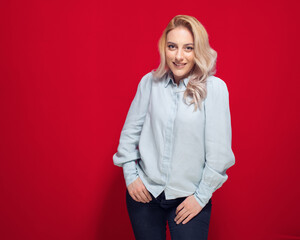 Lovely beautiful young woman smiling while standing with hands in pockets, isolated on red background. Half length of girl with happy laugh.