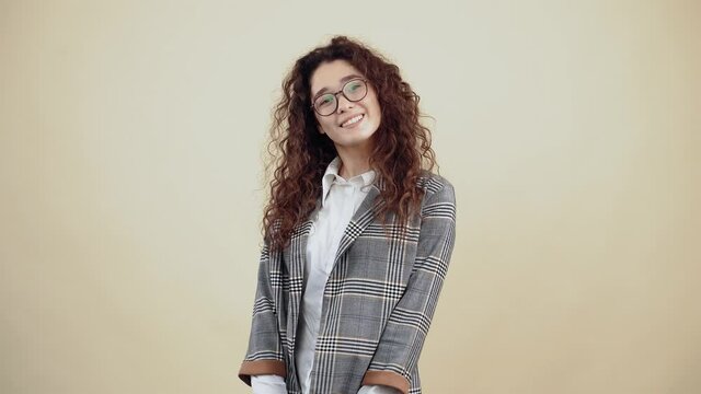 Portrait of a young woman with curly hair in a gray jacket and white shirt, with glasses posing isolated on a beige background in the studio. The concept of people's lifestyle. Smile wide and