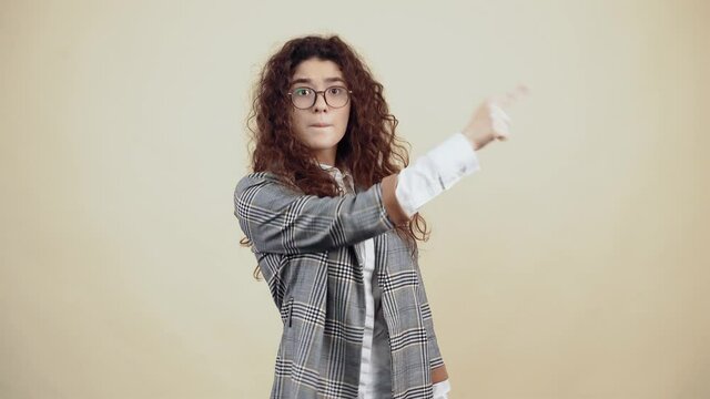 The surprised young woman, shocked with her curly hair in a gray jacket and white shirt, with glasses posing isolated on a beige background in the studio. The concept of people's lifestyle. Point your