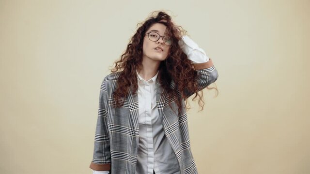 The nice young woman arranges her hair, looking in the room Cretaceous in gray jacket and white shirt, with glasses posing isolated on a beige background in the studio. The concept of people's
