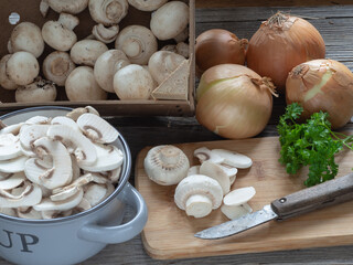 Raw mushrooms, whole and sliced, lined from a wooden board box and placed in a metal pot. Light onions and parsley leaves.