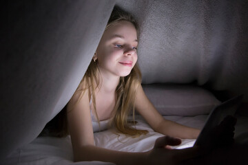 Obraz premium child, cute girl, listening to music and playing games on a tablet, at night under a blanket, hiding from parents