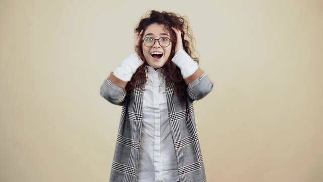 Shocked, excited young woman with her hands in her hair says wow looking curiously at the camera Cretaceous in gray jacket and white shirt, with glasses posing isolated on a beige background in the